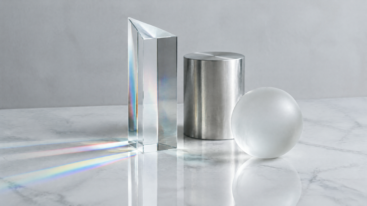 Glass prism refracting subtle bands of colored light across a white marble surface in a pale grey studio