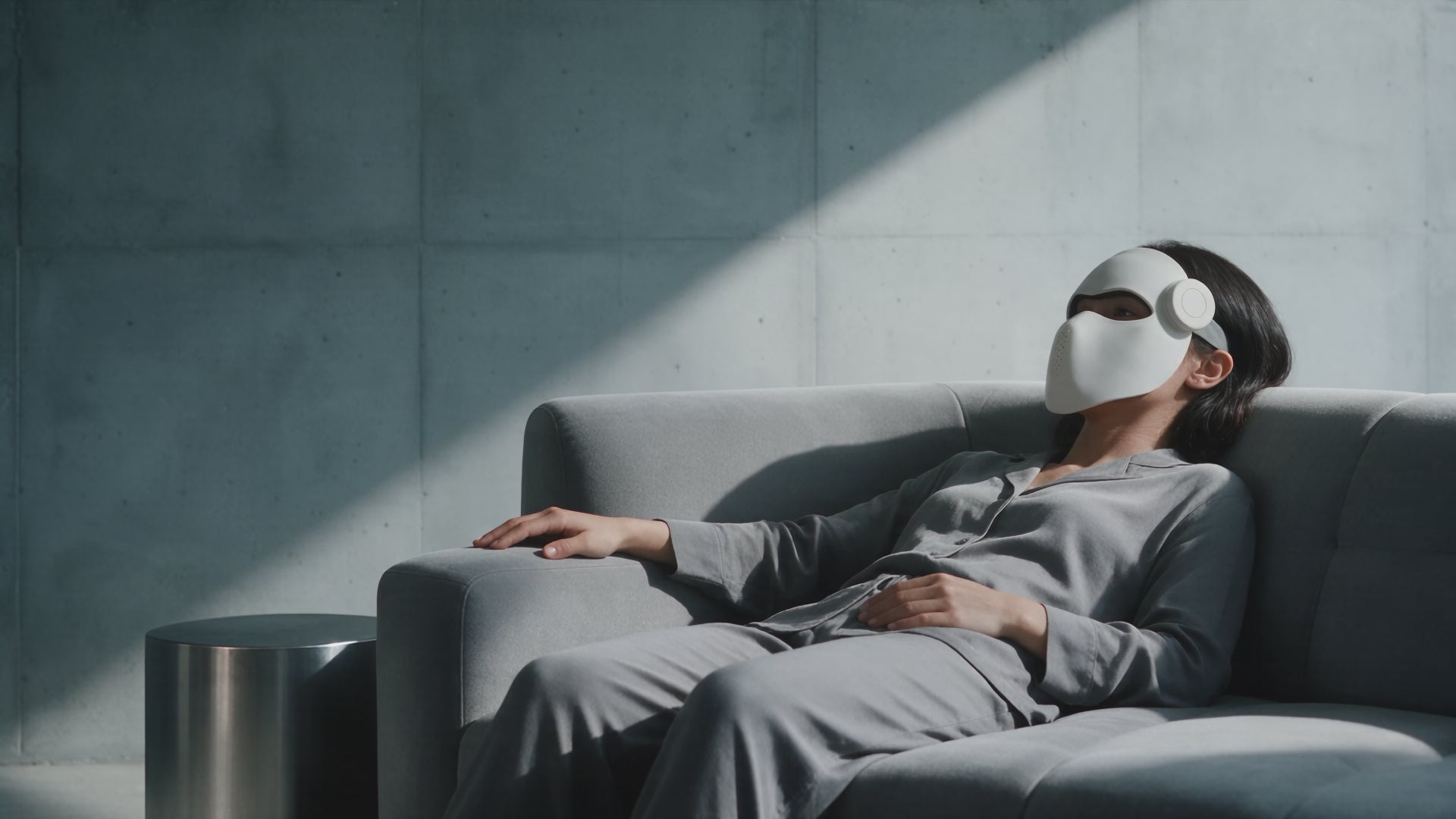 A woman relaxing on a gray sofa while wearing the white Luxen Veil mask